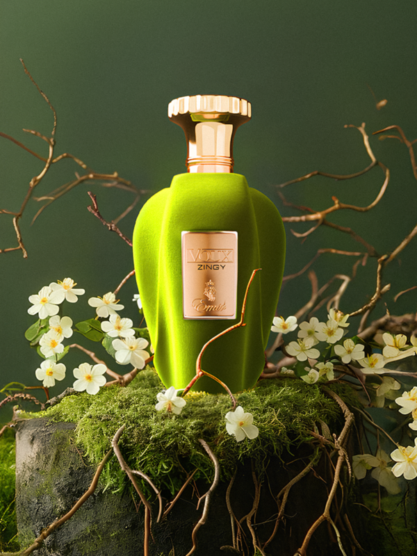 Perfume Paris Corner Voux Zingy en frasco verde con tapón dorado rodeado de flores blancas y ramas sobre fondo verde