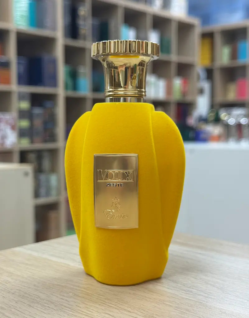 Frasco del perfume Voux Zeste en color amarillo con detalles dorados sobre mesa en interior de tienda de perfumes