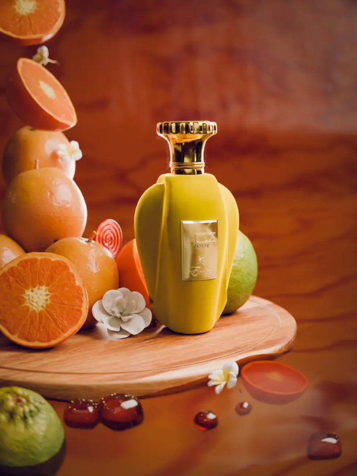 Perfume Voux Zeste en frasco amarillo rodeado de naranjas, cítricos y elementos dulces sobre fondo cálido